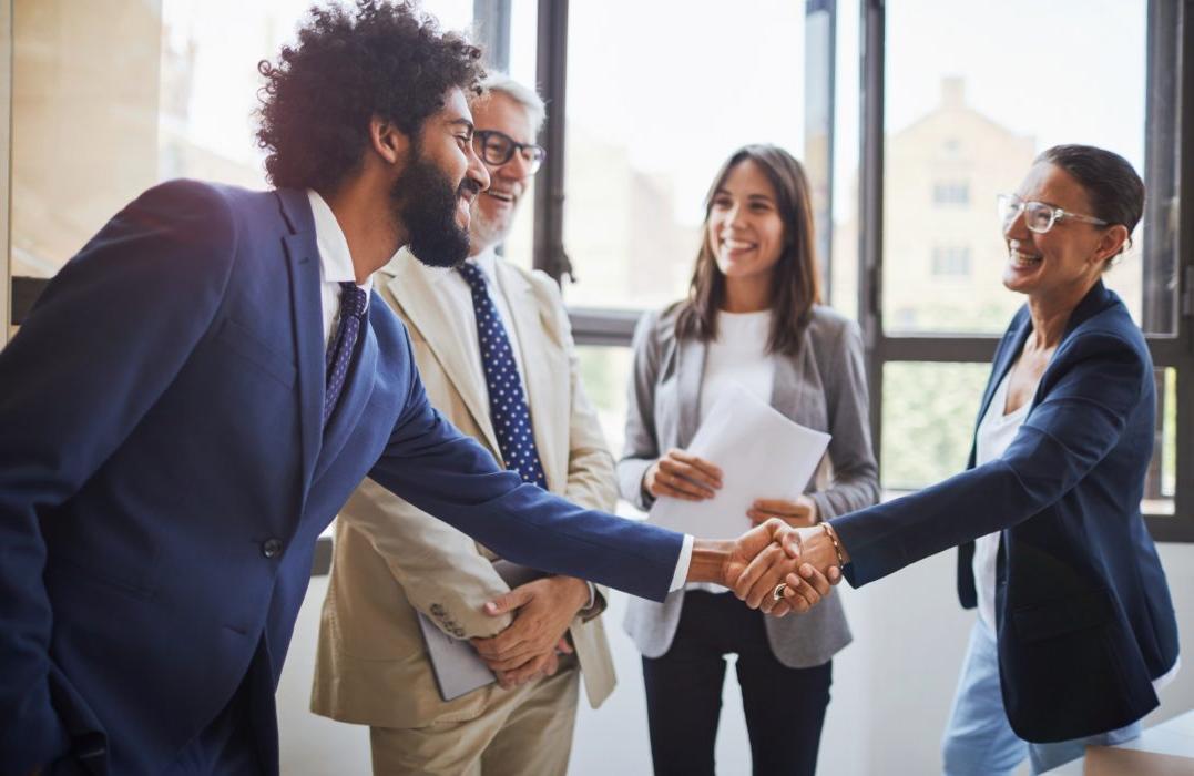 Four people engaging in conversation, two of whom are shaking hands. There are three men and one woman in this photo.